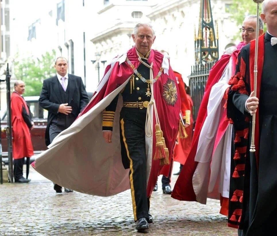 Un homme âgé en uniforme à galons dorés, collier et plaque, marche dans un manteau rouge doublé de blanc. Autour de lui, des personnes en robes rouges et noires, l'une tenant une masse.