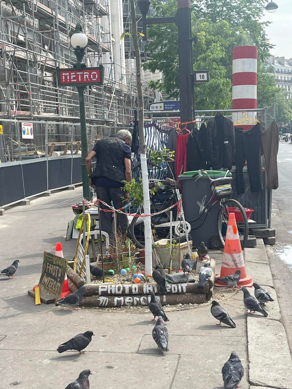 Un homme se penche près d'un arbre sur un trottoir : vélo, poubelle, vêtements sur cintres, pigeons partout. Panneau Métro, échafaudages et inscription « Photo interdit merci ».