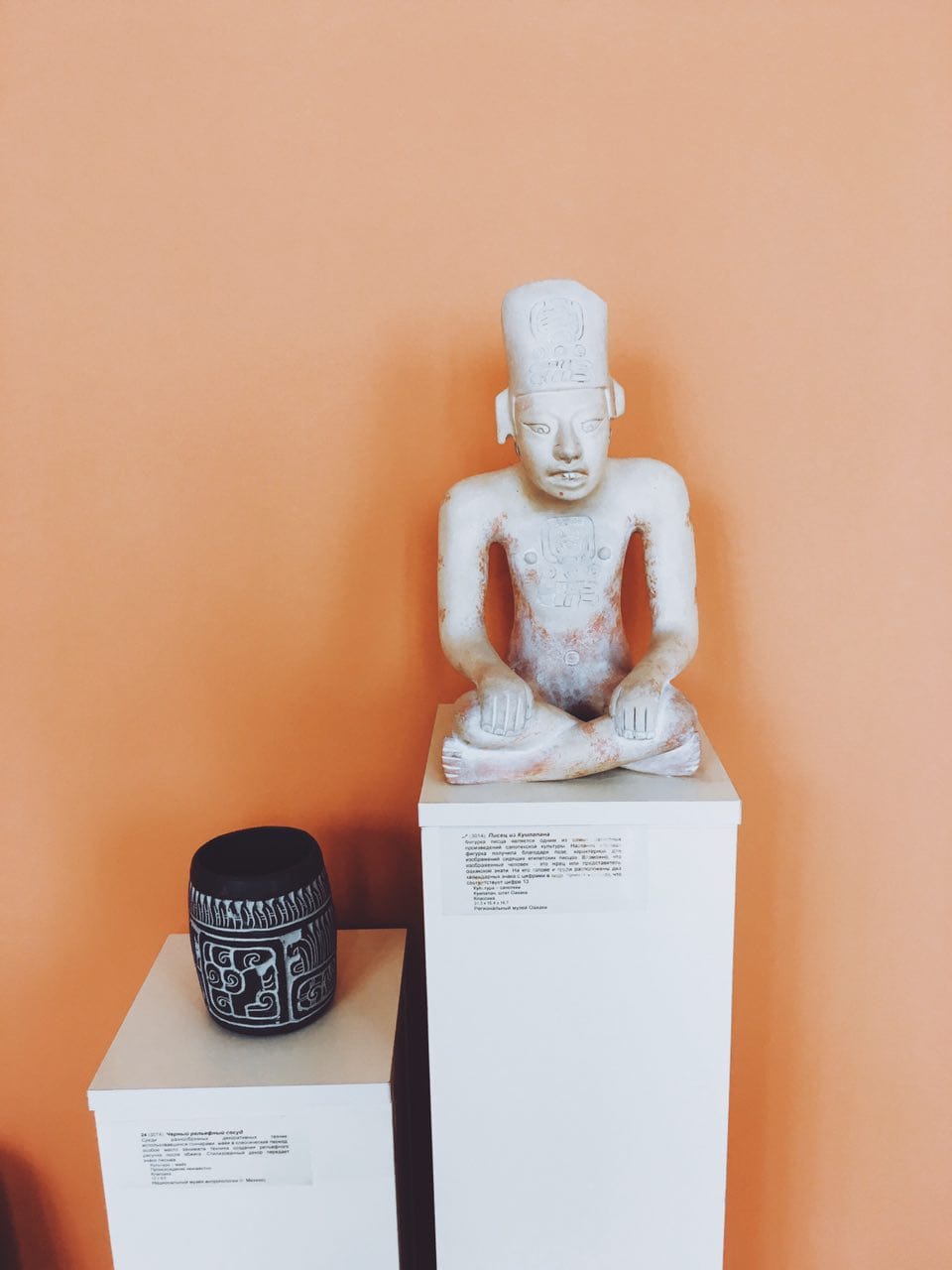 Figurine blanche d'un scribe assis en tailleur sur un socle blanc, près d'un vase noir à décor en relief, devant un mur orange.