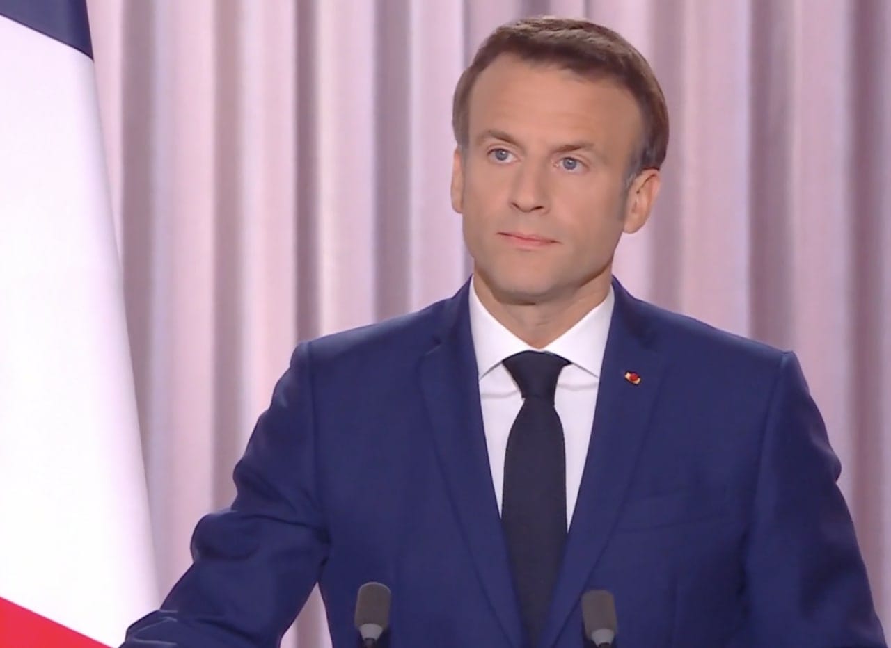 Un homme en costume bleu foncé et cravate noire se tient devant des microphones près d'un drapeau français, devant des rideaux roses.