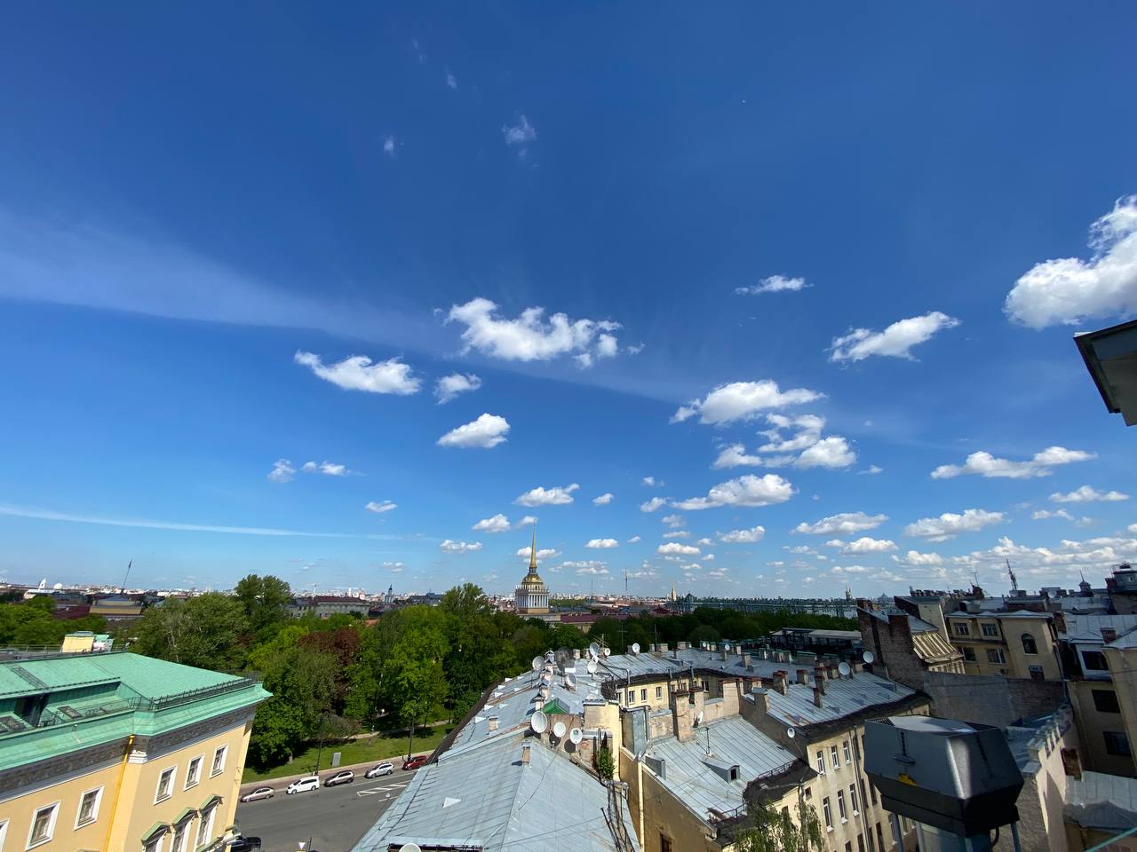 Toits avec antennes paraboliques au premier plan, arbres verts et flèche dorée de l'Amirauté à l'horizon sous un ciel bleu parsemé de nuages.