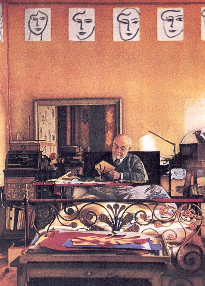 Elderly Henri Matisse in glasses sits in bed working with coloured paper, with a row of drawn faces on the wall above him.
