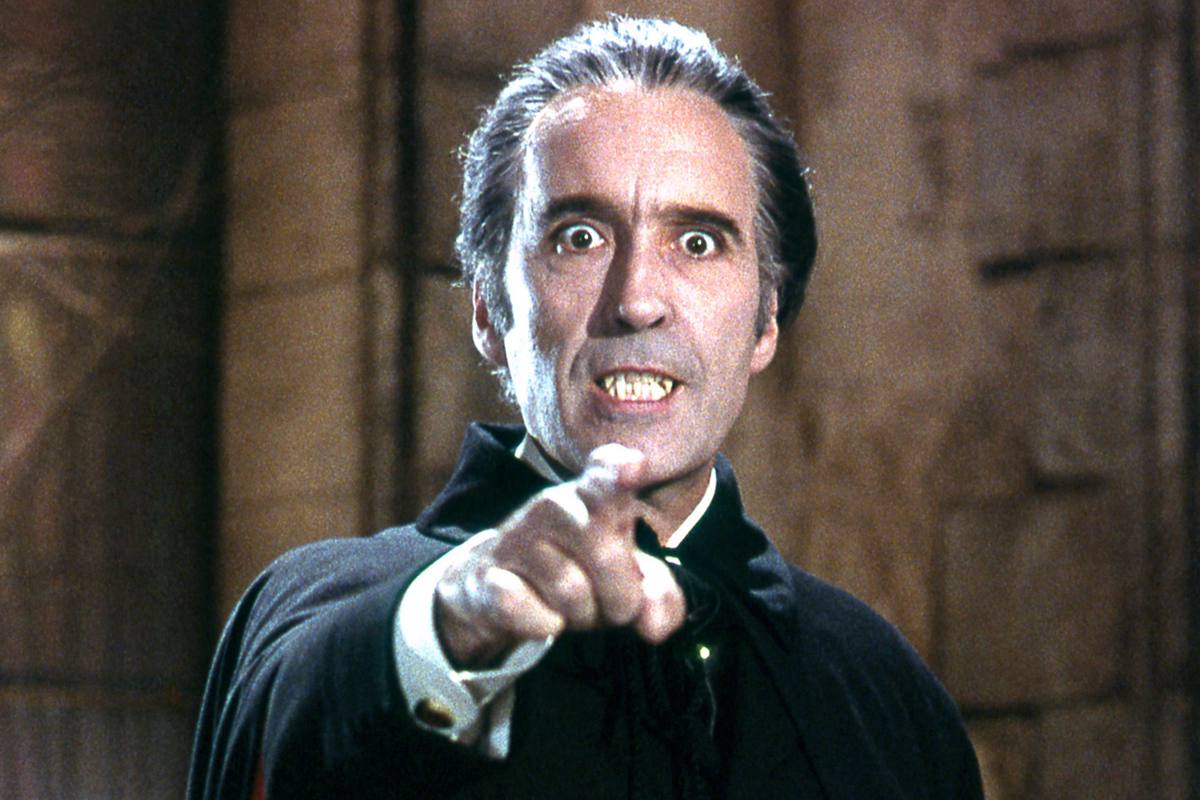 A man in a dark cloak with slicked-back hair points at the viewer, with fangs visible in his mouth.