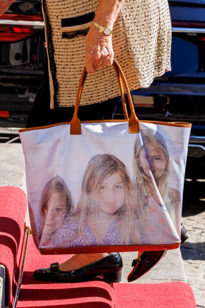 Une femme âgée en veste de tweed tient un grand sac aux anses brunes orné d'une photo de trois fillettes. Un tapis rouge et des chaussures vernies noires sont visibles.