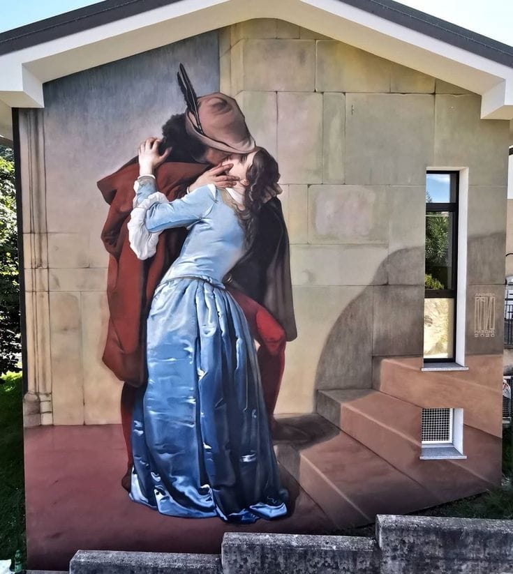 Fresque sur le pignon d'un bâtiment : un homme en chapeau à plume et cape rouge embrasse une femme en robe de satin bleu. Marches et ombre peintes se fondent dans le mur.