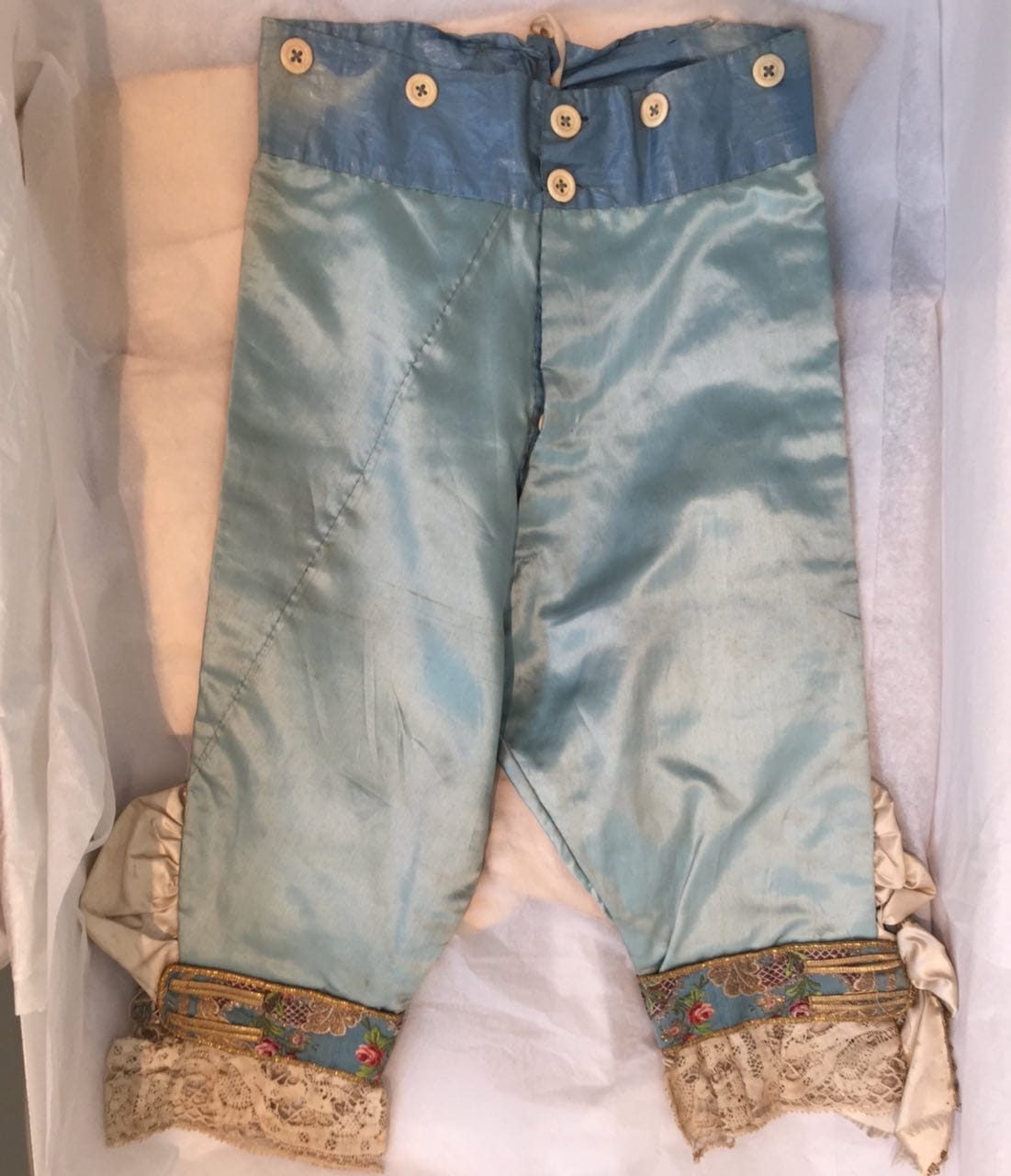 Culottes de satin bleu clair, avec boutons à la ceinture, dentelle et bas brodés, posées dans une boîte sur du papier de soie blanc.