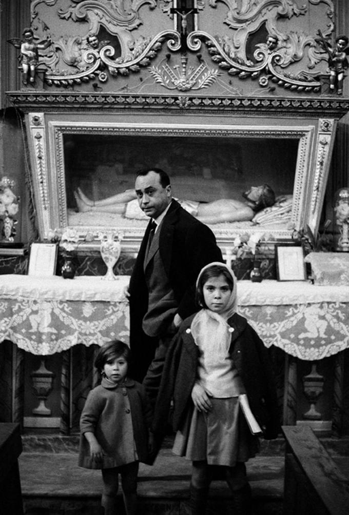 Photographie en noir et blanc : un homme en manteau et deux fillettes, l'une coiffée d'un fichu, posent dans une église devant un autel orné où repose un Christ gisant, sous une nappe de den