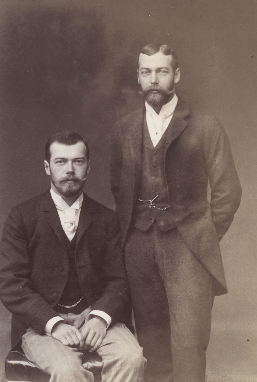 Old sepia photograph of a seated young bearded man in a suit and a taller bearded man standing beside him with a hand behind his back.