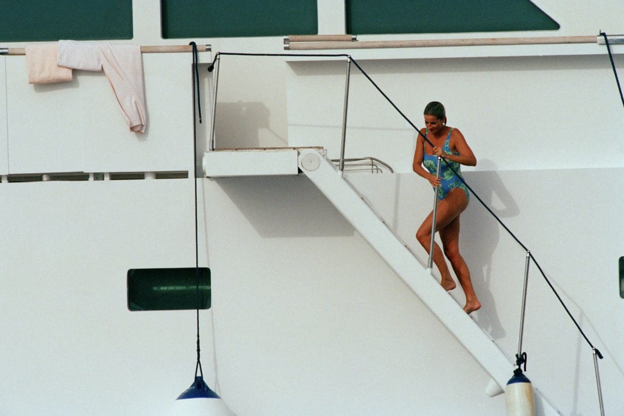 Une femme en maillot bleu à motif vert descend un étroit escalier le long de la coque blanche d'un navire. Des serviettes sèchent à gauche ; des bouées pendent en bas.