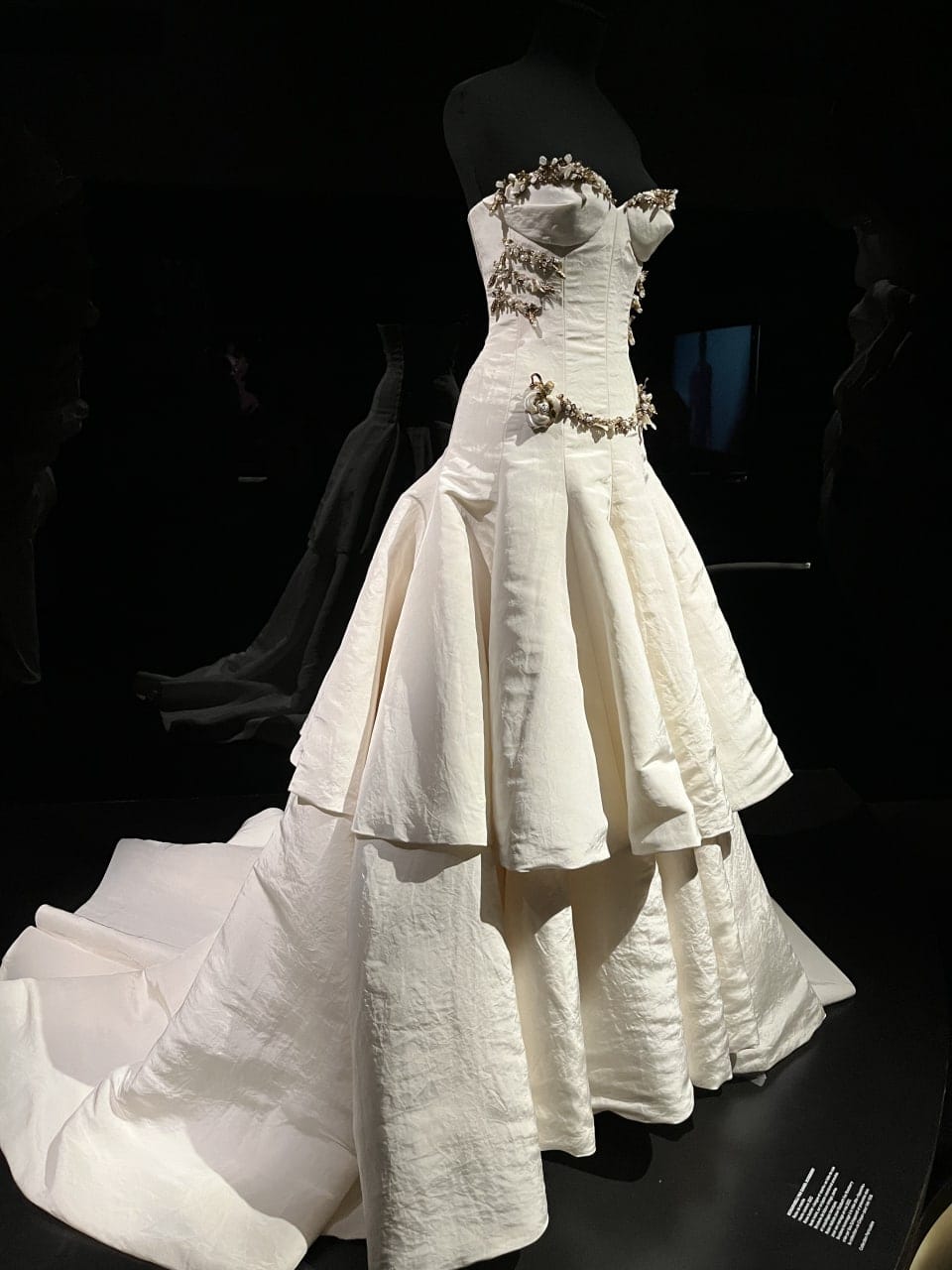 Robe blanc crème au corset brodé de motifs de branches, à jupe étagée et traîne, sur un mannequin noir dans une salle d'exposition sombre.