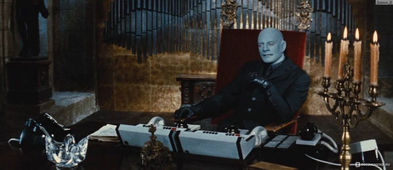 Image de film : un homme au crâne bleu et chauve, ganté de noir, est assis à un bureau avec des téléphones, près d'un candélabre allumé.