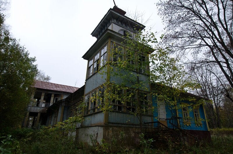 Maison en bois abandonnée avec une haute tour, des fenêtres brisées et une véranda, entourée de buissons envahissants et d'arbres nus sous un ciel couvert.