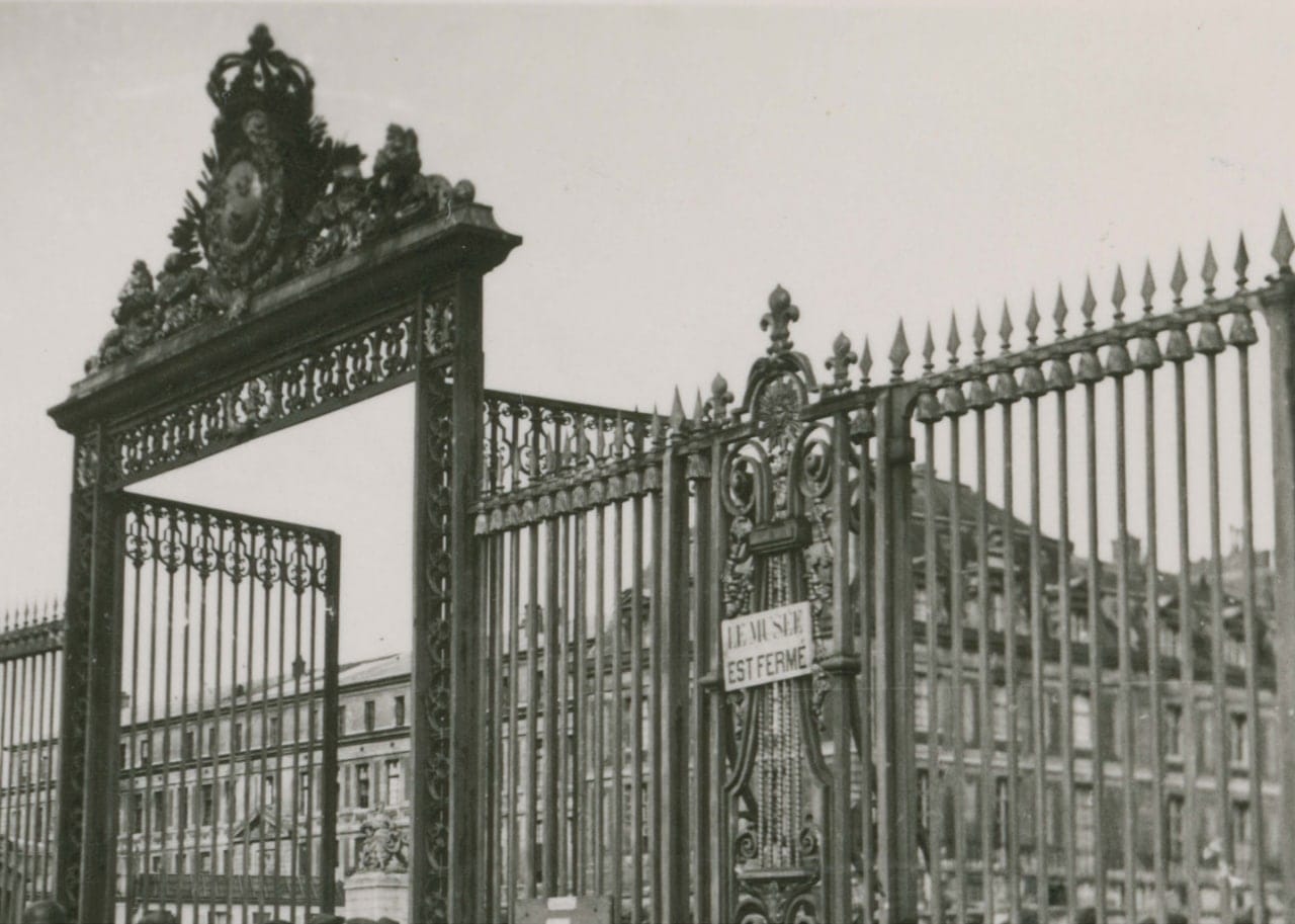 Photographie en noir et blanc des grilles en fer forgé de Versailles, surmontées d'une couronne, avec l'écriteau « Le musée est fermé ».