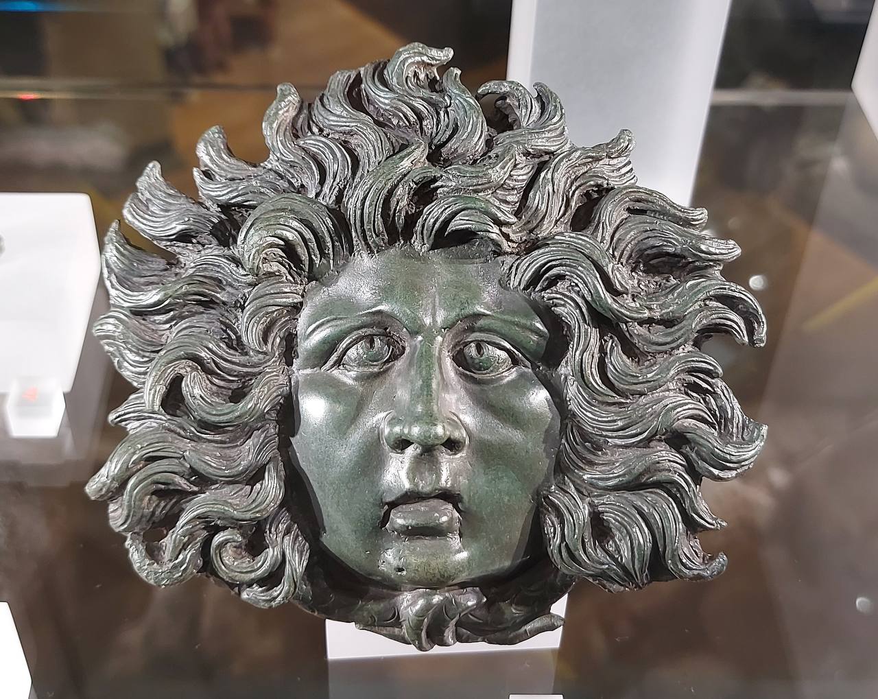 Relief head of Medusa Gorgon with a startled face and thick, wavy locks of hair, displayed in a museum case.