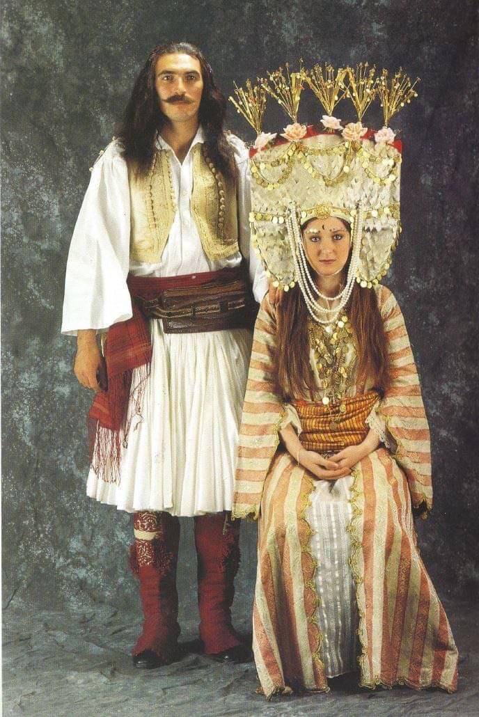Portrait en studio : un homme moustachu aux cheveux longs, en chemise blanche, fustanelle et guêtres rouges, se tient près d'une femme assise en robe rayée et haute coiffe ornée.