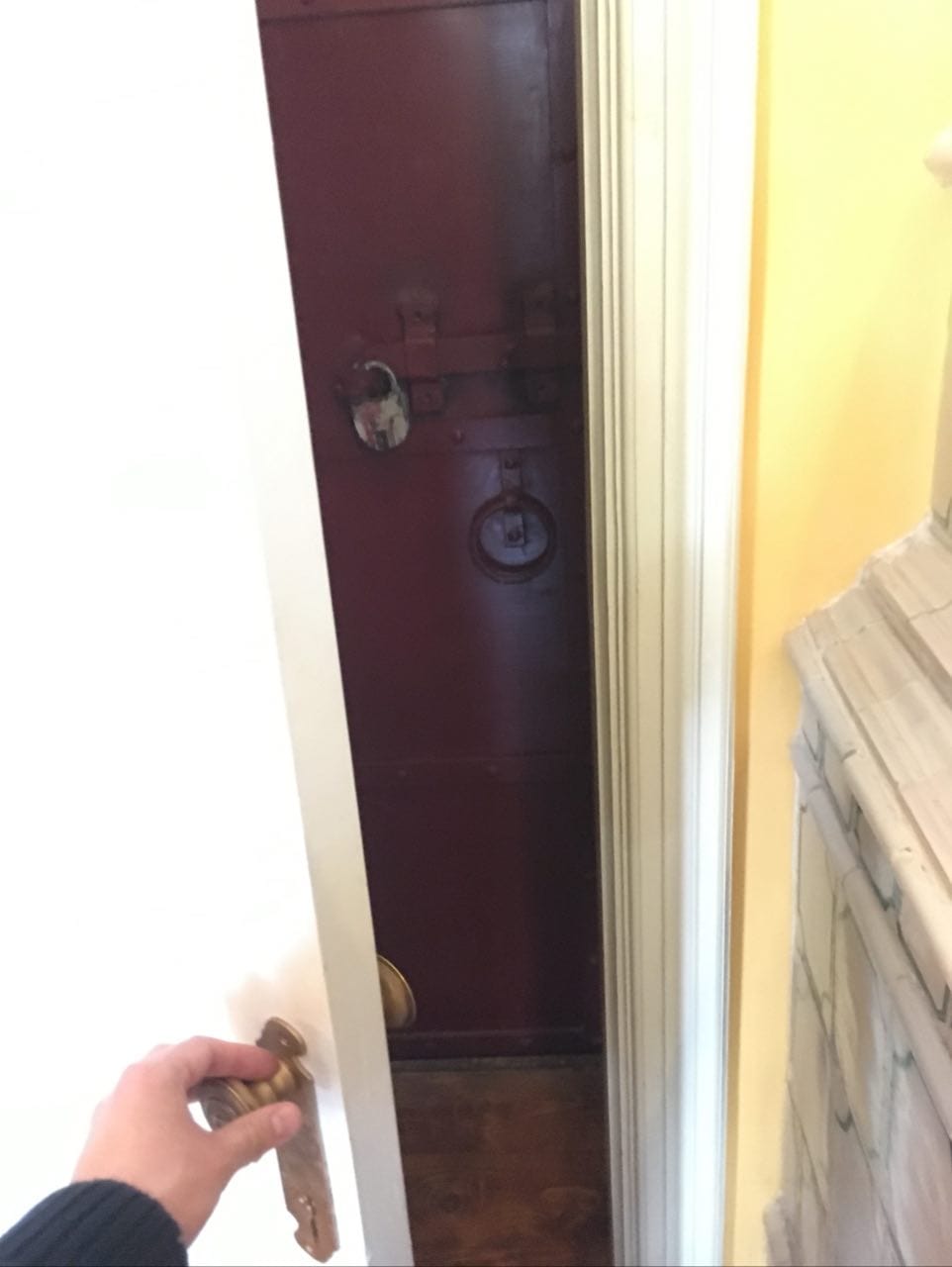 A hand opens a white door, revealing a dark red iron door with a padlock, a bolt and a ring handle.