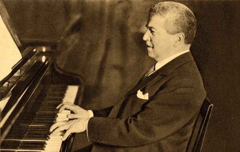 A man in a dark suit with a white pocket square plays a grand piano, shown in profile in a sepia photograph.