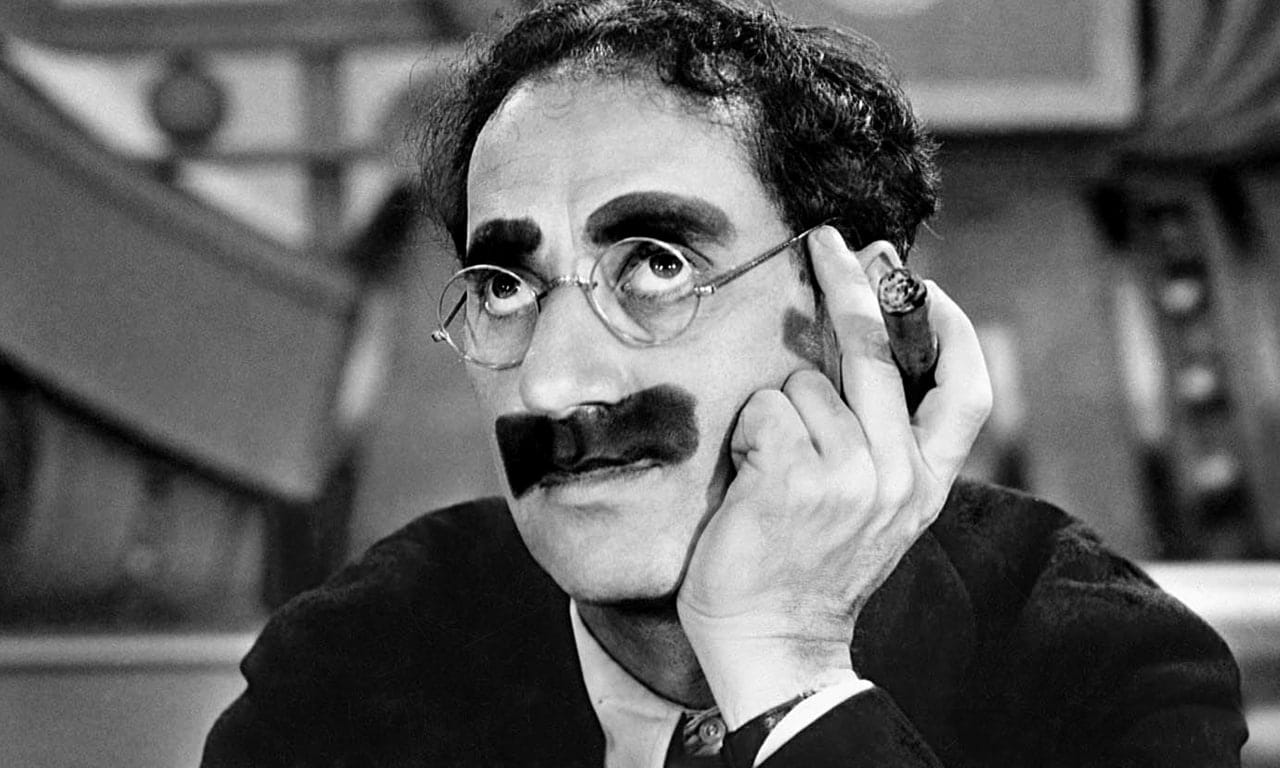 Portrait en noir et blanc d'un homme à lunettes rondes, aux sourcils et à la moustache épais et sombres, tenant un cigare près de la joue.
