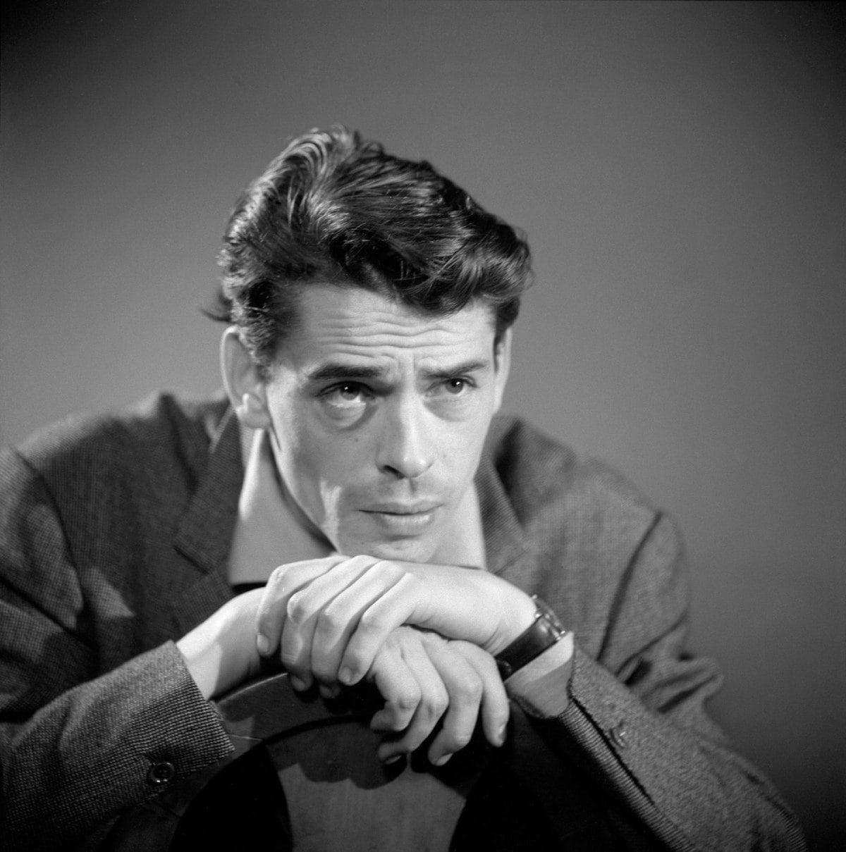 Black-and-white portrait of Jacques Brel in a jacket, chin resting on folded hands, gazing thoughtfully to the side.
