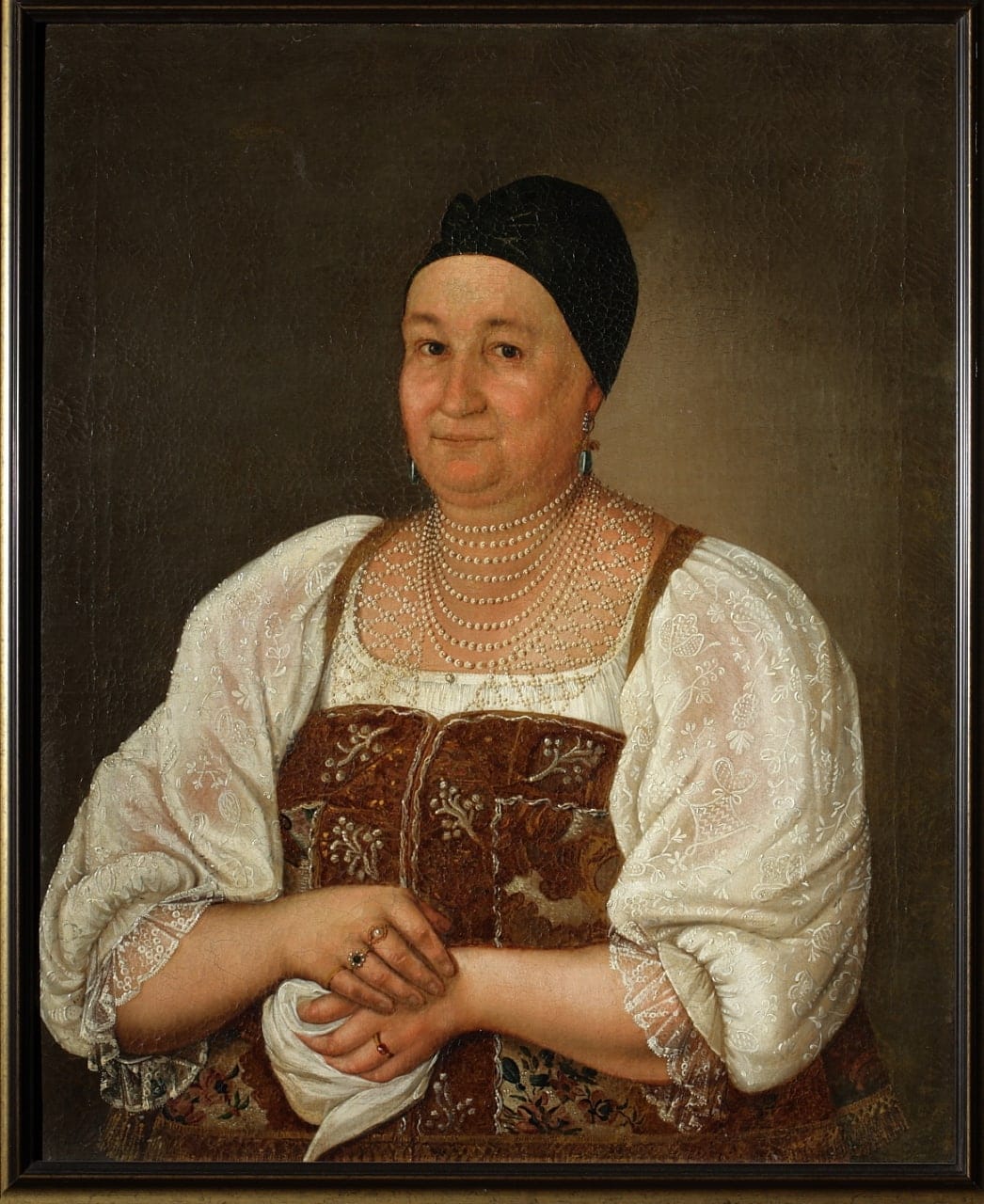 Portrait peint d'une femme âgée coiffée d'un bonnet sombre, en chemise blanche brodée et sarafan de brocart. Elle porte un collier de perles à plusieurs rangs et des bagues, les mains jointe