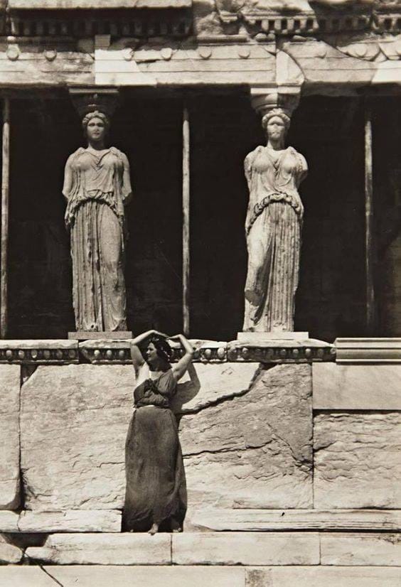 Black and white photograph of a woman in a long dress standing with her arms raised above her head beneath two caryatid statues of an ancient temple.