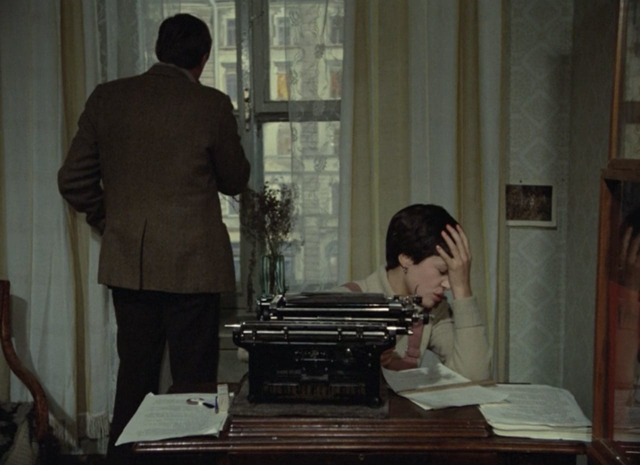 A man in a jacket stands with his back to the viewer at a window, while a woman at a desk with a typewriter and papers rests her head in her hand.