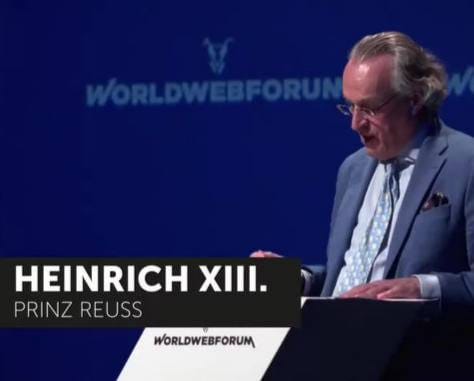 Un homme à lunettes, cheveux gris rejetés en arrière, veste bleue et cravate à motifs, à un pupitre devant un fond bleu World Web Forum ; légende : Heinrich XIII. Prinz Reuss.