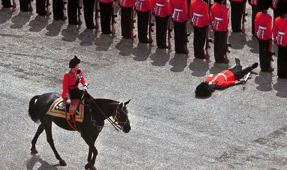 A soldier in a red tunic lies on the ground in front of the ranks while a rider in red uniform passes on a dark horse.