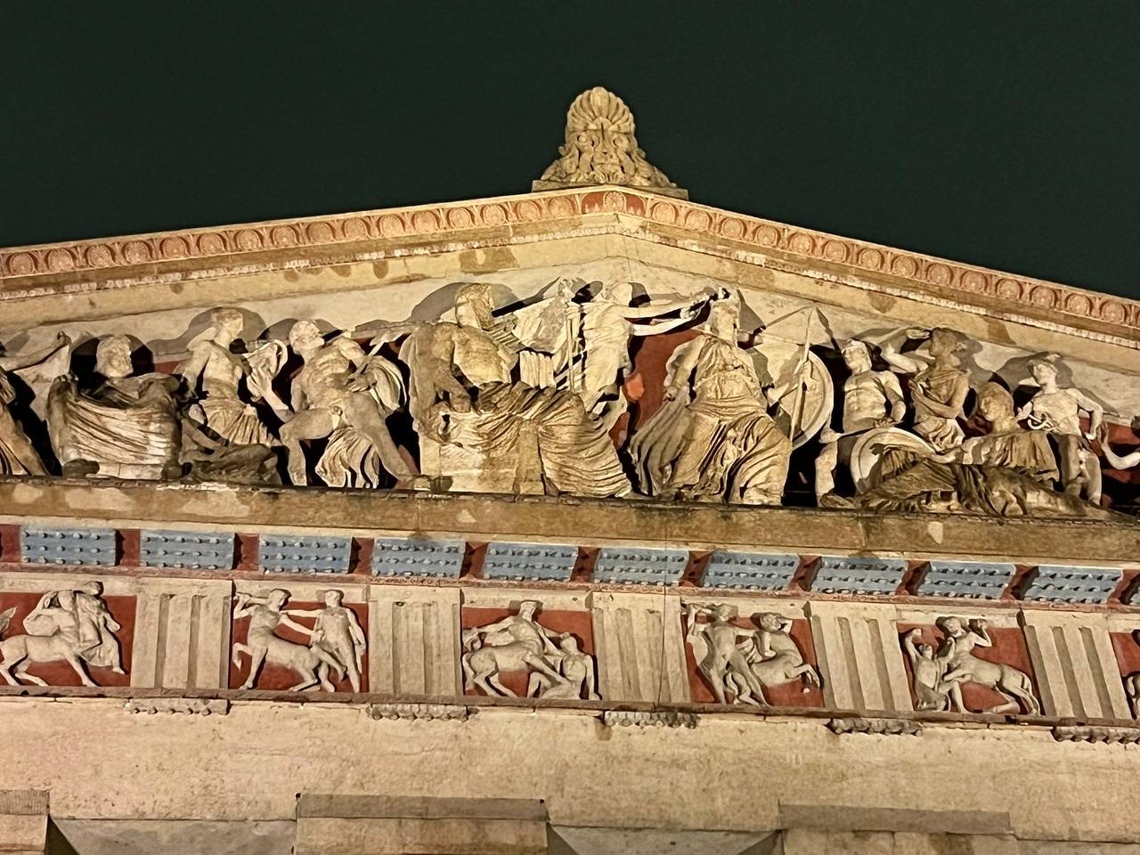 Fronton triangulaire d'un édifice néoclassique avec un groupe de figures sculptées ; dessous, une frise de centaures sur fond rouge et des panneaux bleus, sous un ciel nocturne.