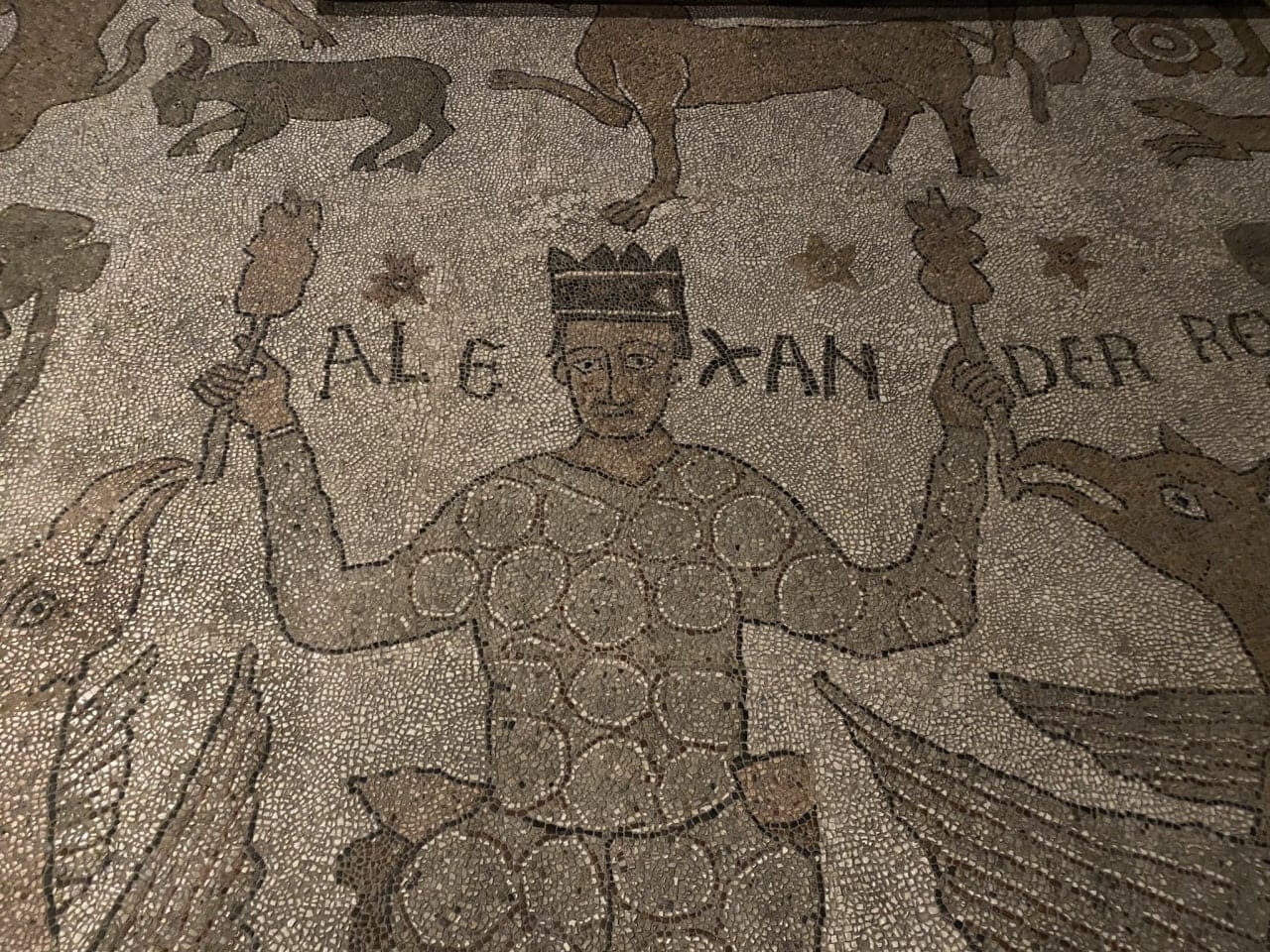 Floor mosaic detail showing a crowned Alexander holding skewers in raised hands between two griffins, with an inscription beside him.
