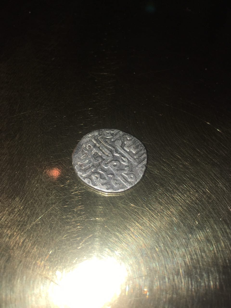 A darkened silvery coin with a relief inscription lies on a dark metal surface with glare.