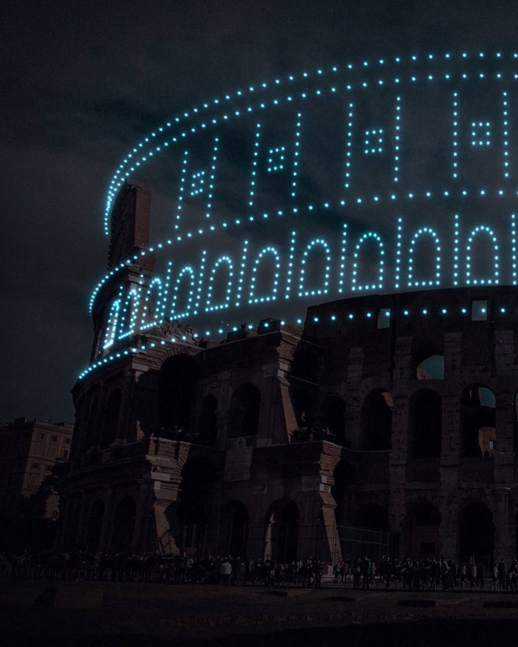 Les ruines sombres du Colisée, avec des points lumineux bleus dans le ciel nocturne qui redessinent arcades et étages. Une foule se tient près de la grille en bas.