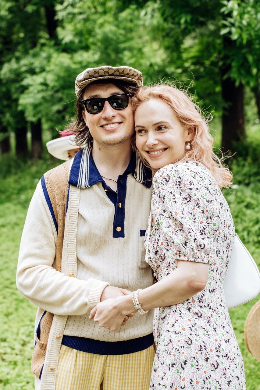 A smiling man in a cap and sunglasses embraces a smiling woman in a floral dress against a green park.