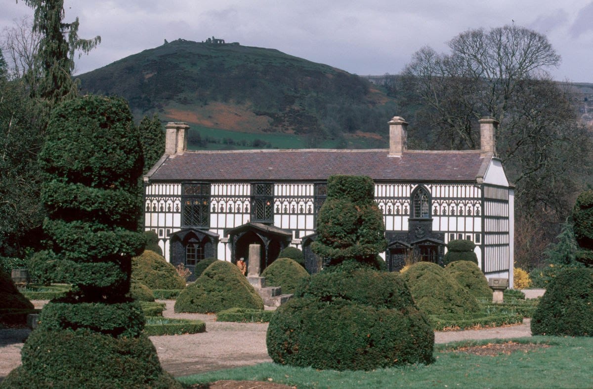 A two-storey black-and-white timber-framed house with clipped topiary shrubs in front and a wooded hill behind.