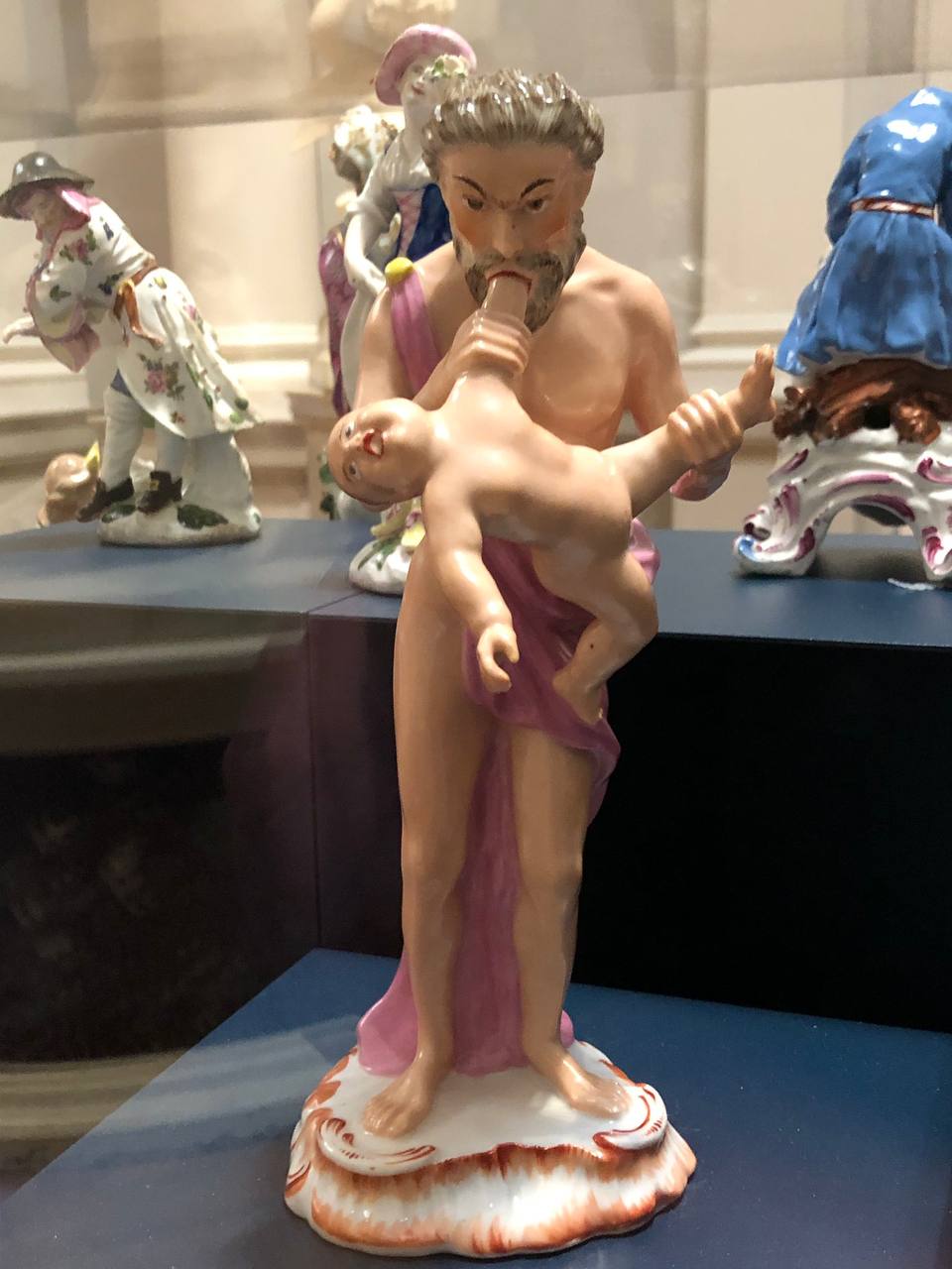 Porcelain figure of a bearded man in pink drapery holding a nude child to his mouth, displayed in a museum case.