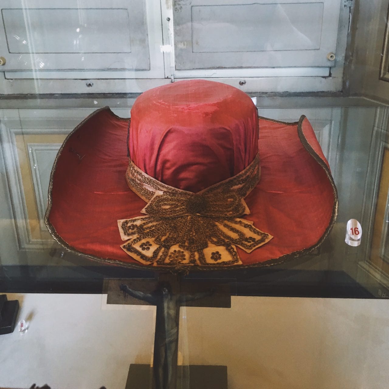 A wide-brimmed red hat with gold embroidery and a bow in a museum display case, with a dark crucifix visible below it.