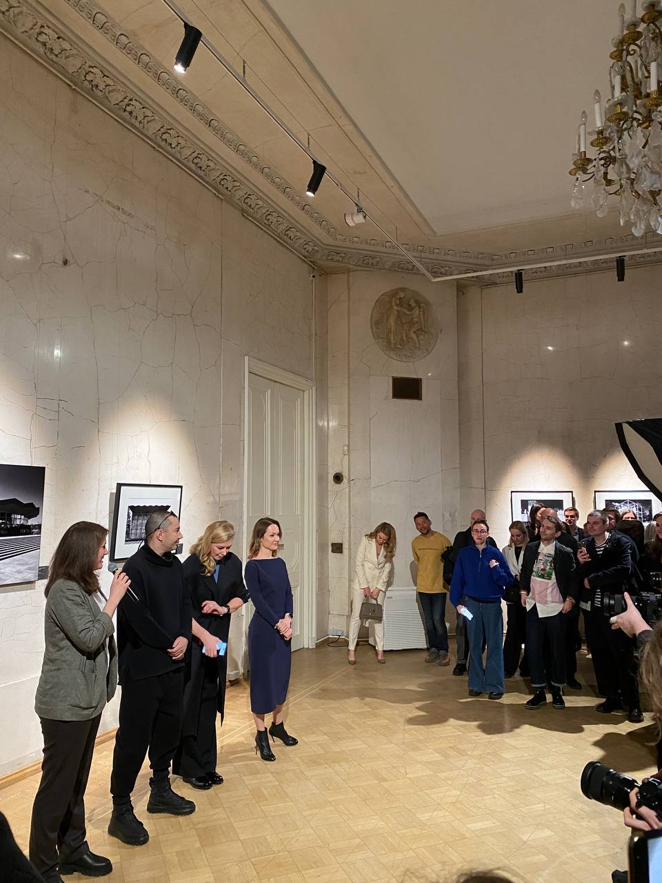 Dans une salle claire à corniche en stuc et lustre en cristal, une femme au micro et trois invités font face au public. Des photos en noir et blanc ornent les murs ; des photographes filment