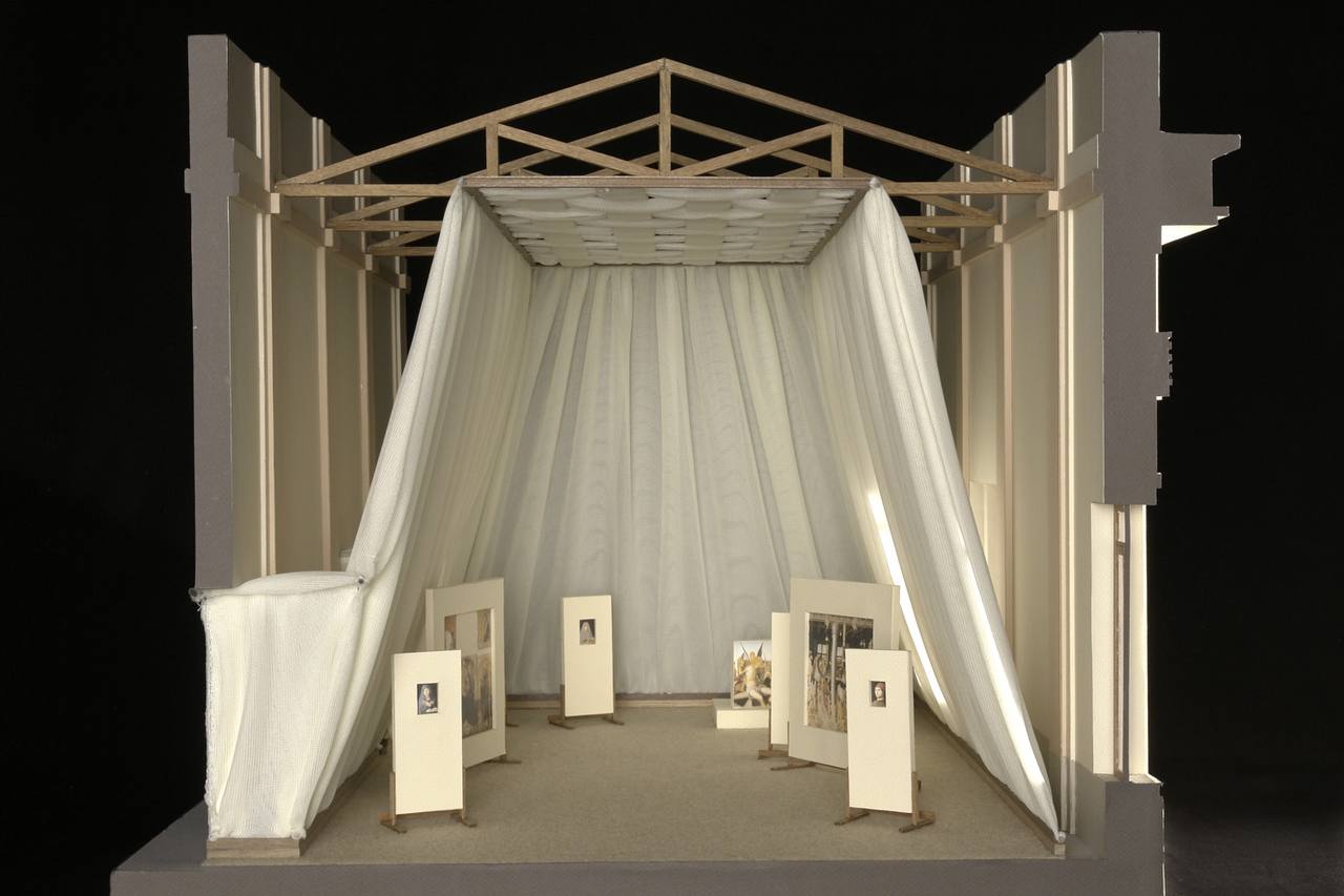 Maquette architecturale en coupe d'une salle à charpente en bois. Un tissu blanc descend du plafond clair vers les murs, et de petits tableaux sont posés sur des supports.