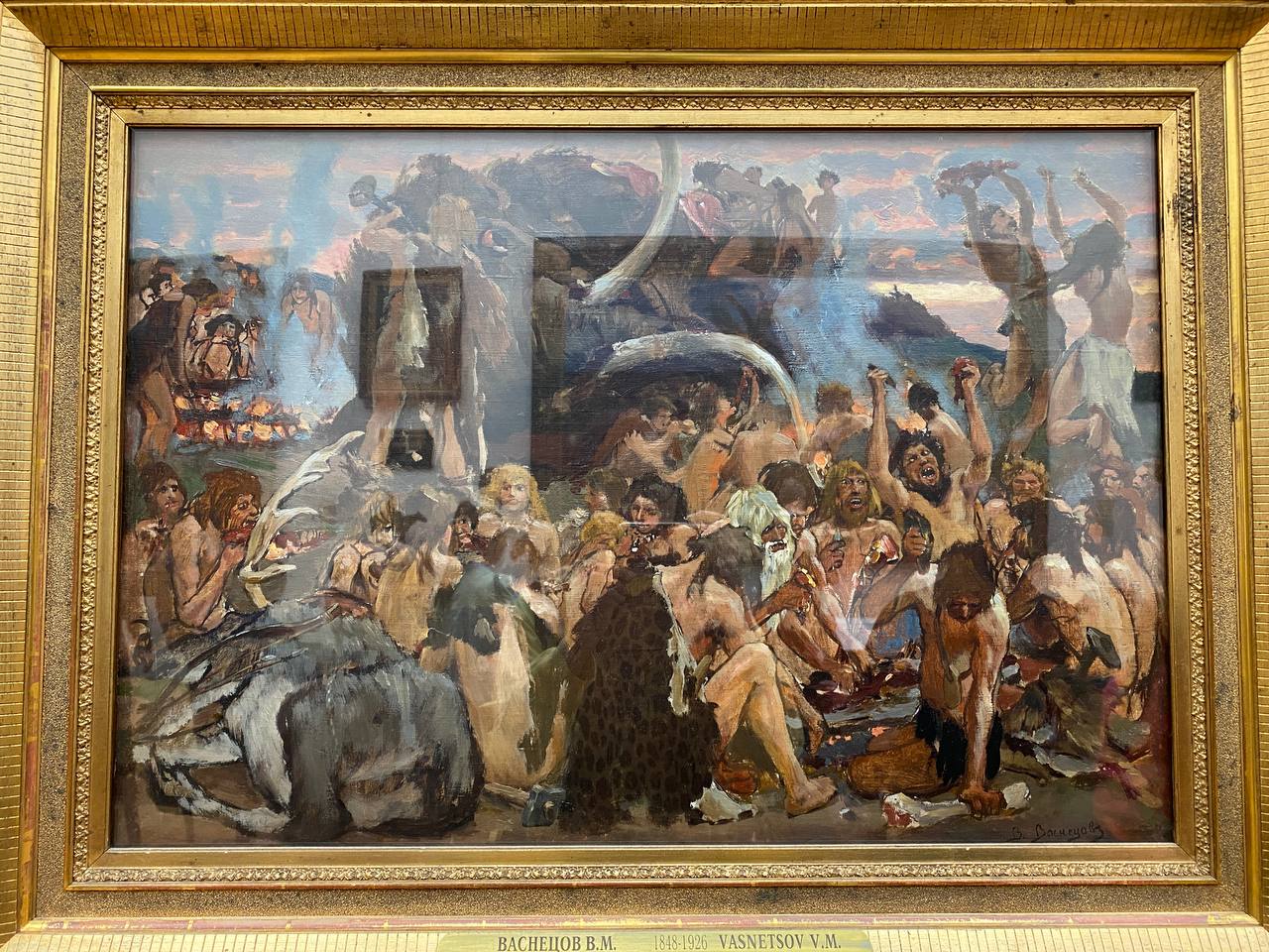 Tableau de Viktor Vasnetsov dans un cadre doré sous verre : des hommes préhistoriques festoient près des feux, un mammouth derrière eux, des reflets sur la vitre.