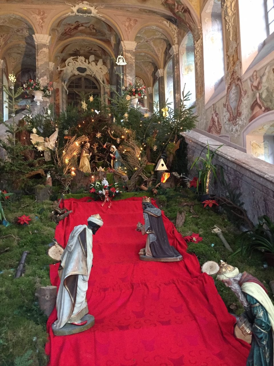 Nativity scene on a staircase in a Baroque interior, with figures of the three kings on red cloth, fir trees and moss, and Mary and Joseph at the top.
