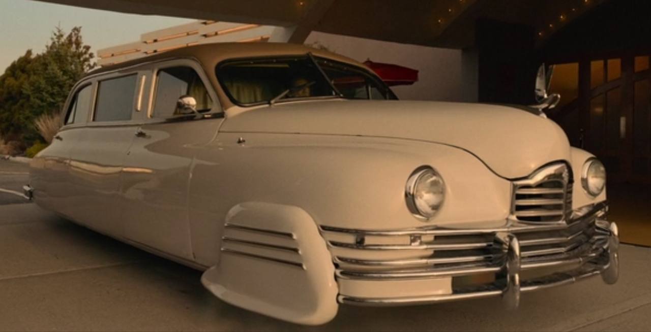 A cream retro-style car with a chrome grille and round headlights stands under a canopy in the light of sunset.