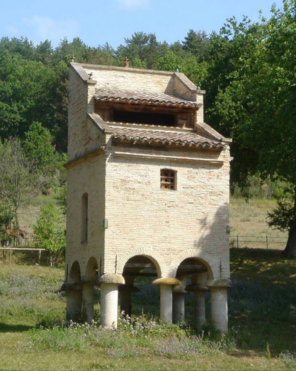 Une tour de pigeonnier en brique claire à toits de tuiles repose sur des piliers ronds à chapeau plat et des arcades. Un pré et une colline boisée l'entourent.