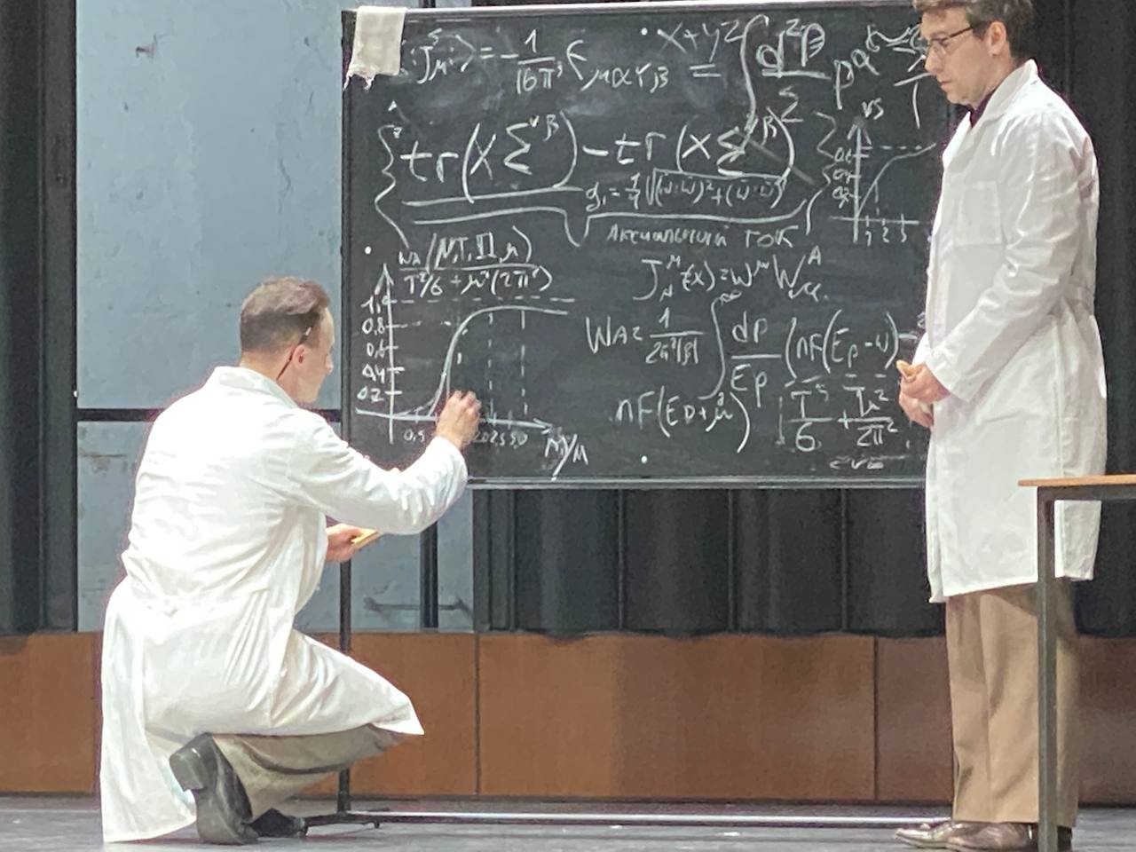 Sur une scène, un homme en blouse blanche accroupi écrit à la craie sur un tableau couvert de formules et de graphiques. À côté, un homme à lunettes, également en blouse, se tient debout.