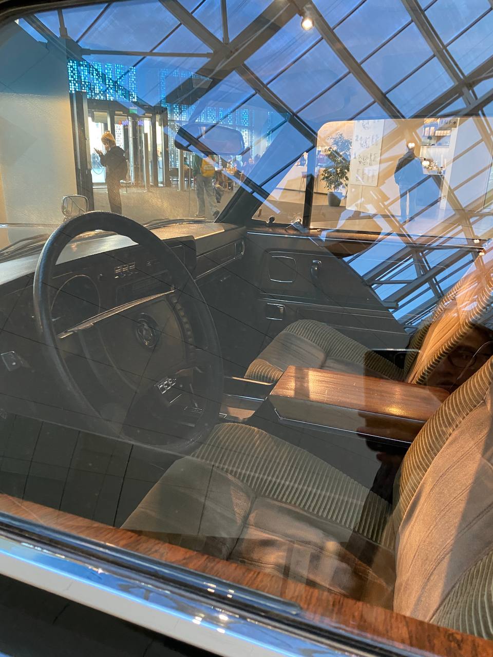 Intérieur d'une voiture ancienne : volant, tableau de bord, sièges en velours côtelé et accoudoir en bois. La vitre reflète une verrière et des visiteurs.