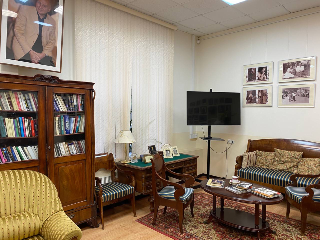 Pièce avec un grand portrait d'homme au mur, une bibliothèque vitrée, un bureau, un canapé et des fauteuils rayés, une table basse et des photographies en noir et blanc.