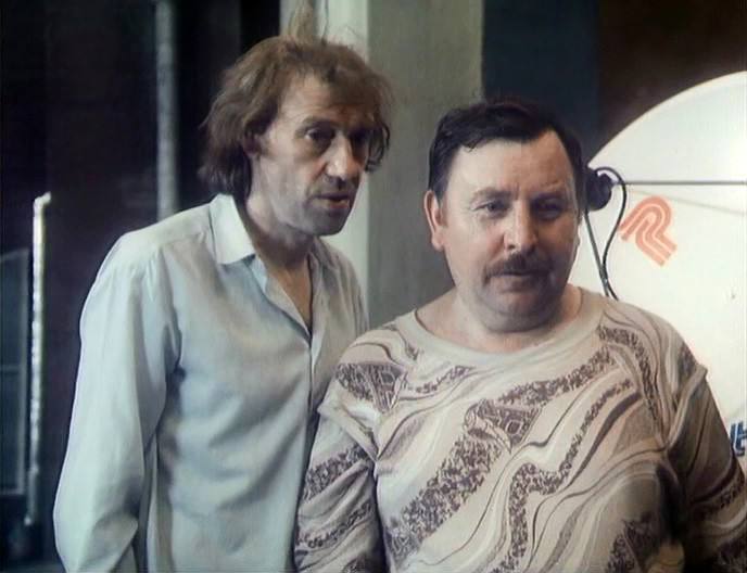 Image de film : un homme maigre aux cheveux ébouriffés, en chemise claire, se tient près d'un homme moustachu en pull à motifs. Une parabole blanche est visible à droite.