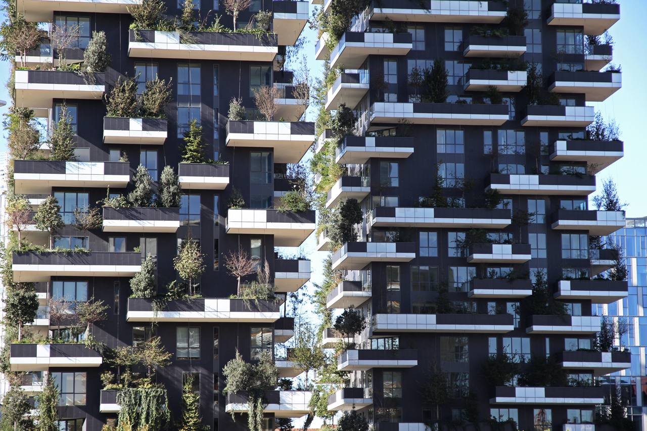 Façades de deux tours sombres. Des arbres et des arbustes poussent sur de larges balcons aux bords blancs, sous un ciel bleu.