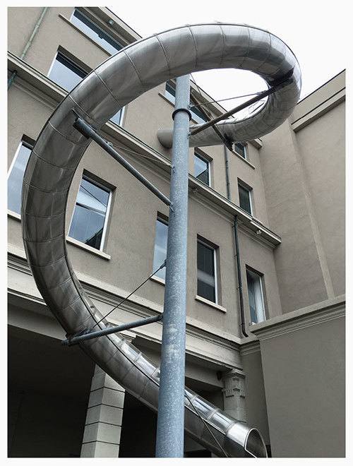 View from below of a spiral metal slide curving along a building wall with windows, with a tall pole in front.