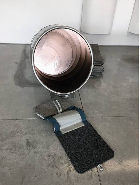 Un cylindre métallique incliné, à l'intérieur brillant, repose sur un sol en béton près de piles de feutre gris et d'un sac en feutre sombre ouvert, à sangles bleues.