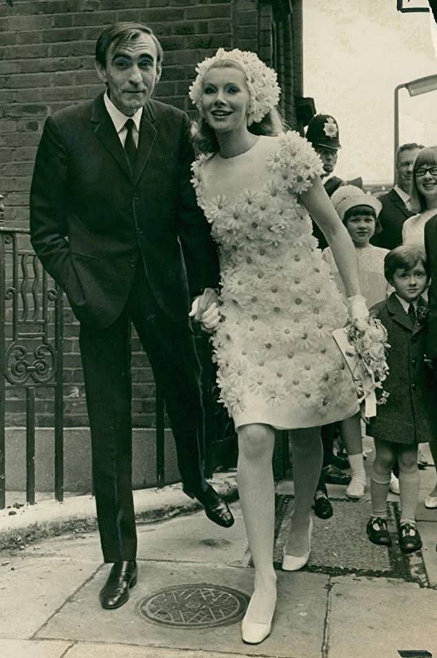 Black-and-white photo of a woman in a short dress and cap covered with flowers walking hand in hand with a man in a dark suit, with children and a policeman behind.